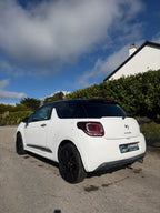 A white Citroèn DS 3 with a black roof and black wheels, parked outdoors on a gravel driveway in front of a white house and green hedges under a blue sky with clouds.