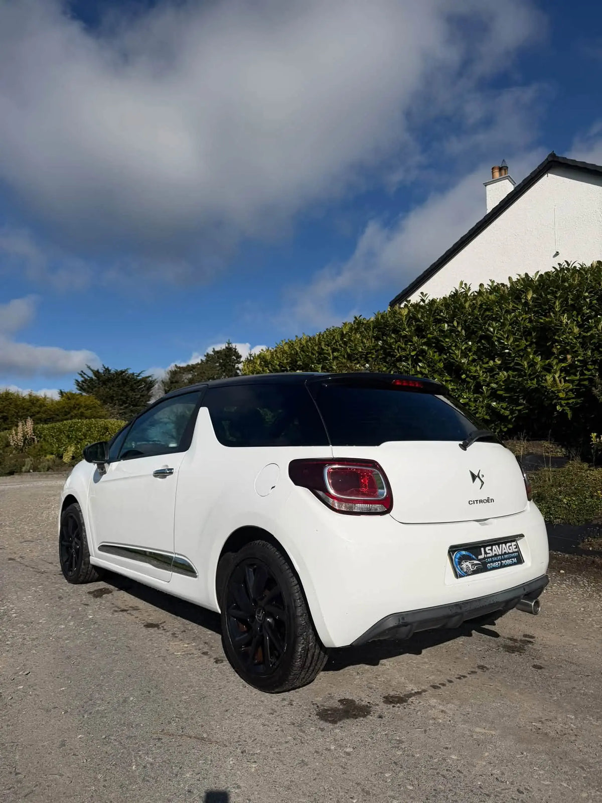 A white Citroèn DS 3 with a black roof and black wheels, parked outdoors on a gravel driveway in front of a white house and green hedges under a blue sky with clouds.