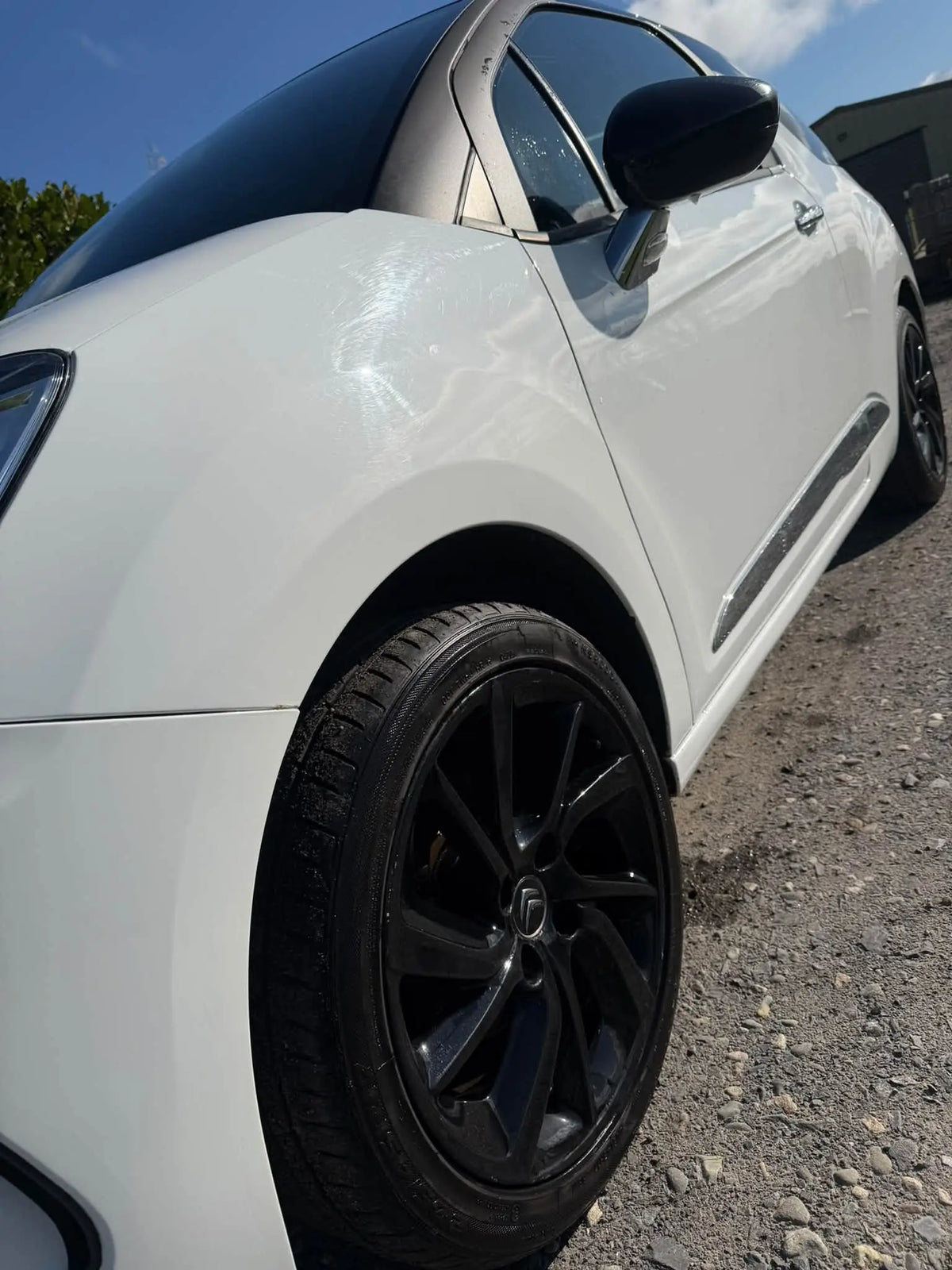 A close-up side view of a white car with black multi-spoke wheels, parked outdoors on a gravel surface under a blue sky.