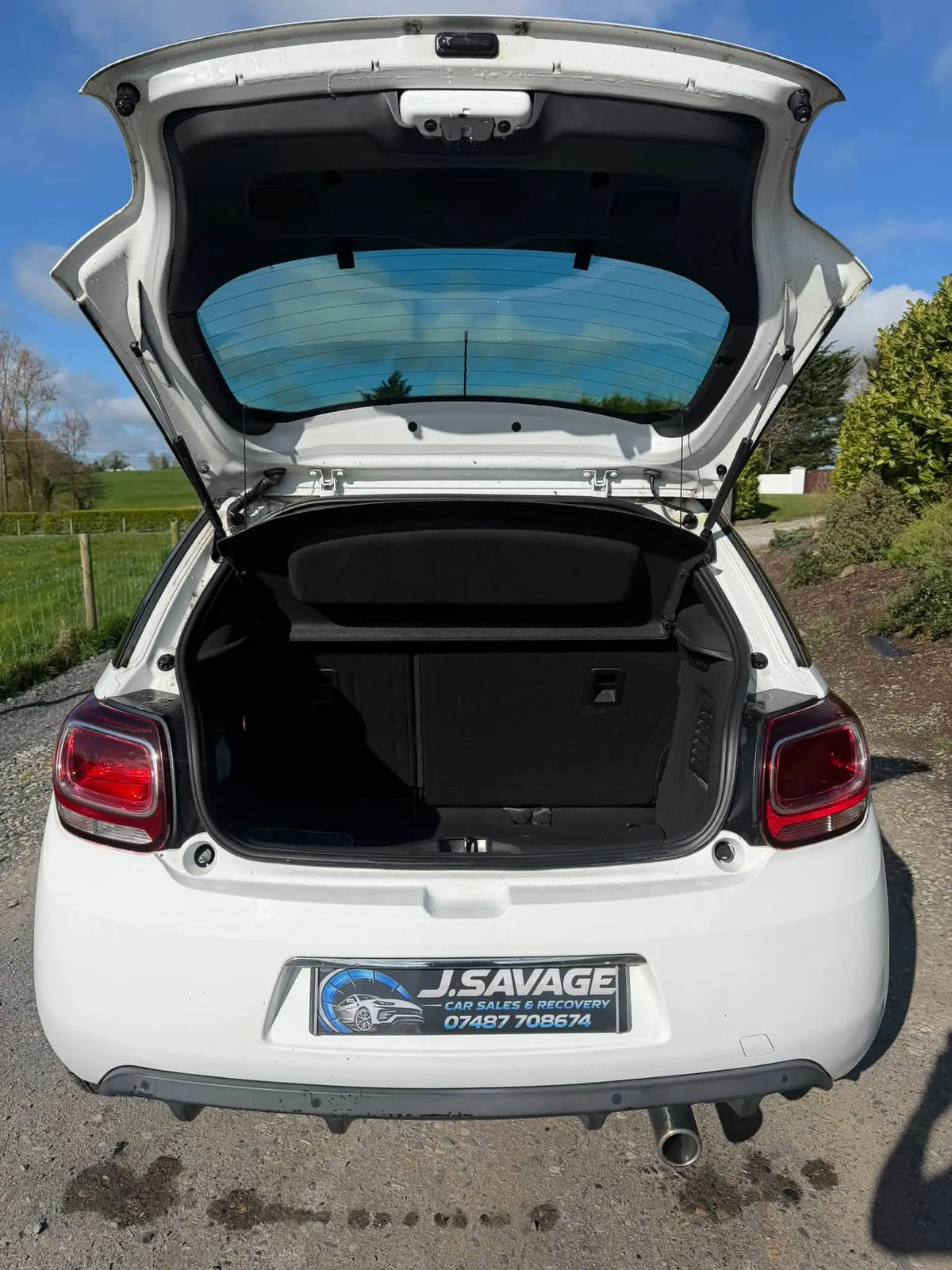 White hatchback car shown from the rear with the trunk open, parked outdoors on a gravel surface with grass, trees, and blue sky in the background.